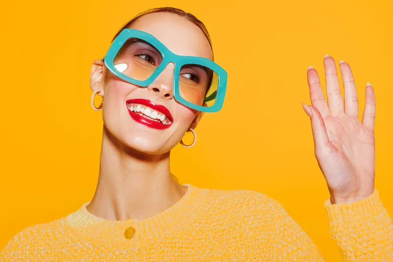 Smiling woman with bright red lipstick and large turquoise glasses waves hand against vibrant yellow background. Concept of fashion, joy, and colorful style in studio portrait.