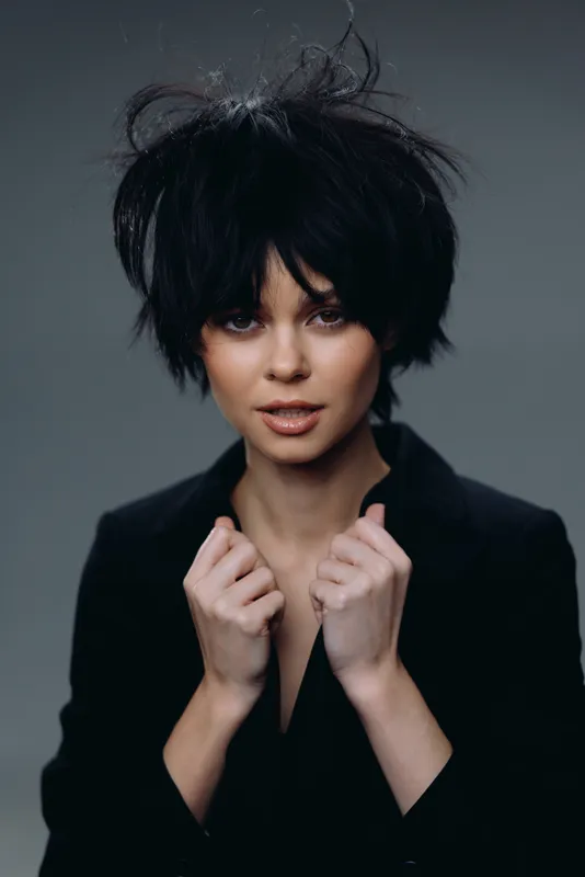Young woman with stylish short black hair wearing a black jacket, posing with confident expression and a dark gray background.