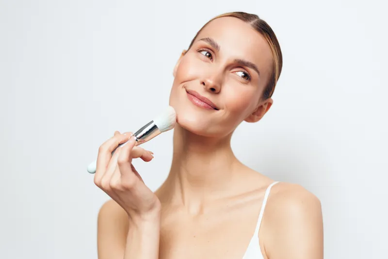 Young woman with clear skin applying powder makeup with a brush on her face. Studio portrait with soft lighting on a white background. Concept of beauty and skincare routine.