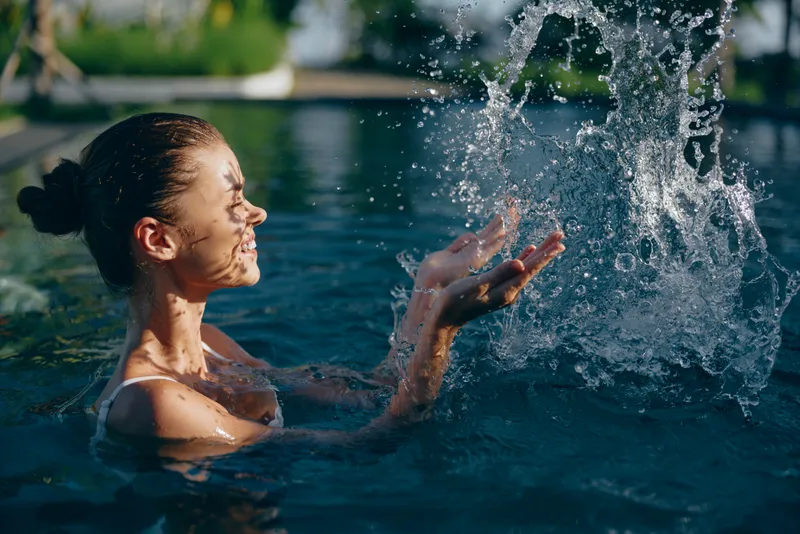 Young woman with brunette hair enjoys playful moment splashing water while swimming outdoors in a sunny natural pool, expressing joy and relaxation in a lifestyle scene.