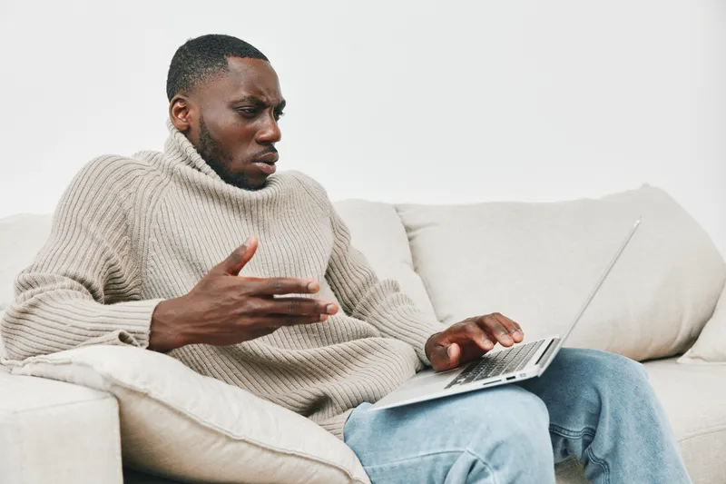 A thoughtful African American man in a cozy beige sweater sitting on a couch, engaging with a laptop, embodying curiosity and contemplation in a minimalist living space