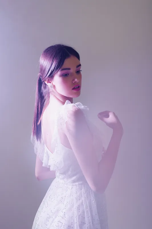 Young woman beauty fashion model in a white lace dress posing with a soft purple light background. Concept for elegance, style, and feminine grace in fashion photography.