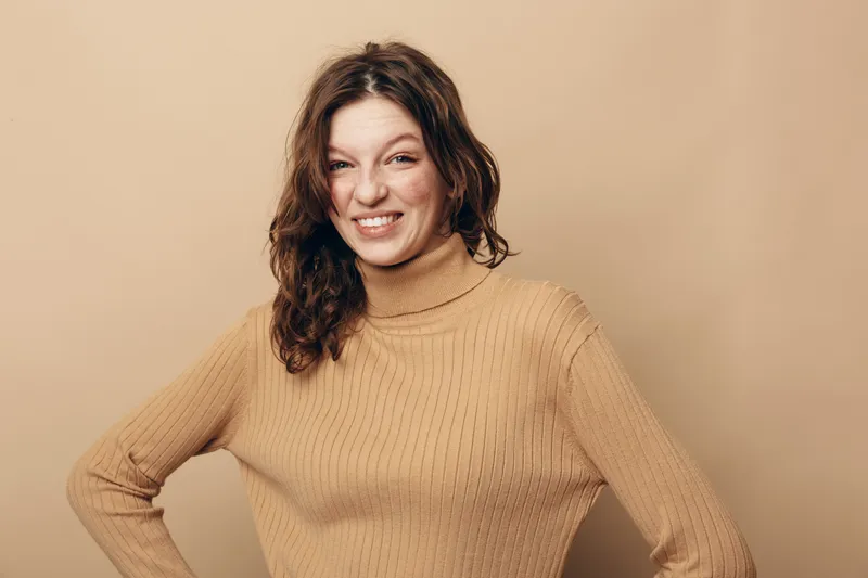 Young woman with curly hair wearing beige turtleneck sweater makes funny face with scrunched nose and squinted eyes. Studio portrait on neutral background with casual style and humor concept.