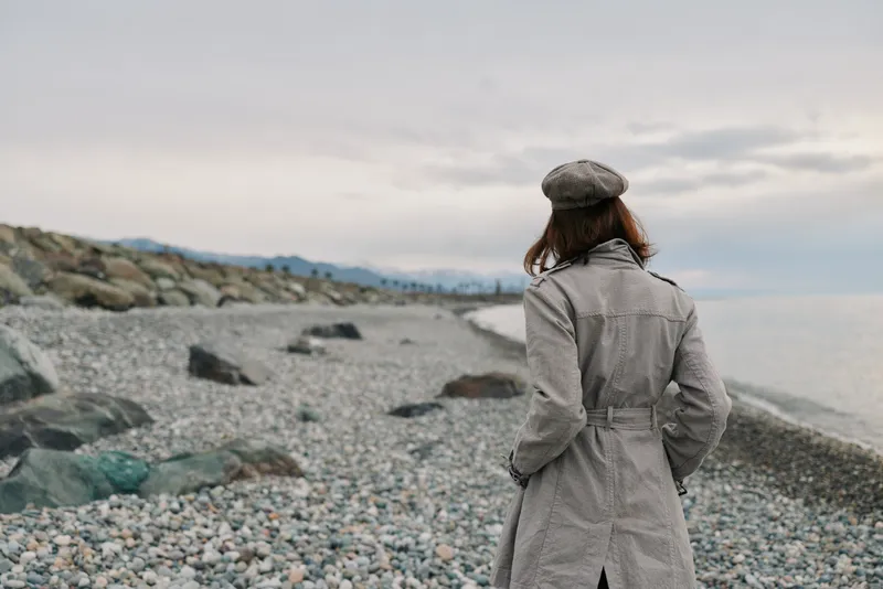 Woman in gray coat and beret walks on rocky beach near calm sea under cloudy sky. Concept of solitude, autumn fashion, and serene outdoor lifestyle by the coast.