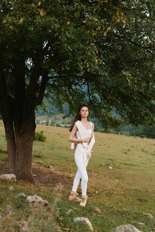 Woman outdoors in a white outfit beside a large tree, relaxed lifestyle moment, natural daylight, open field backdrop, gentle breeze, calm pose, soft colors, candid mood