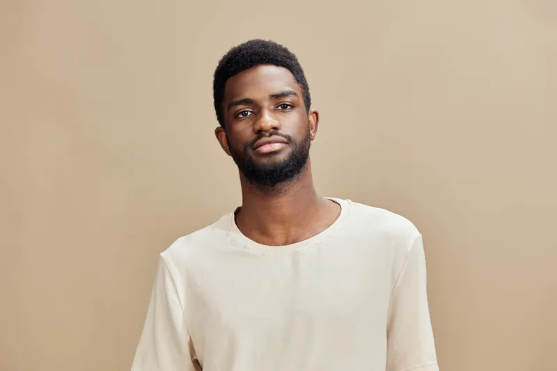 Confident young man in casual beige shirt against a neutral background, showcasing a relaxed yet assertive expression, ideal for lifestyle and fashion concepts