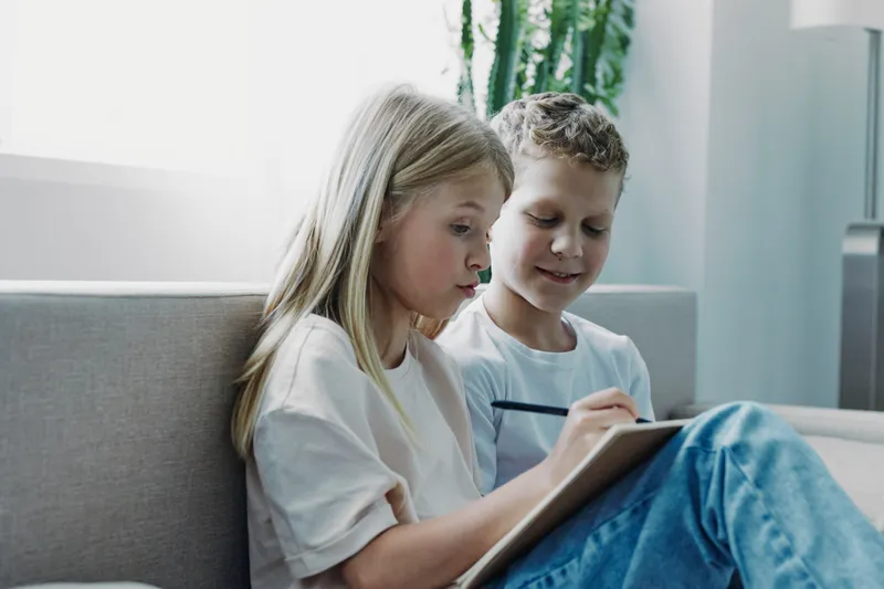 Two children sitting on a sofa focused on writing or drawing on a clipboard in a bright living room. Concept of learning, creativity, childhood, and teamwork in a casual home setting.