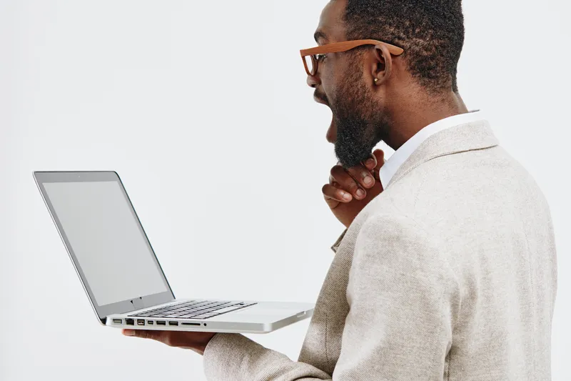 Professional man holding laptop with gray screen, wearing beige blazer and glasses, profile view, standing against plain white background, studio shot, technology and business concept, modern workspac