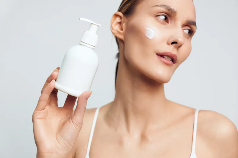 Young woman applying white face cream on cheek while holding a white lotion bottle. Skincare and beauty concept with soft natural lighting and clean white background.