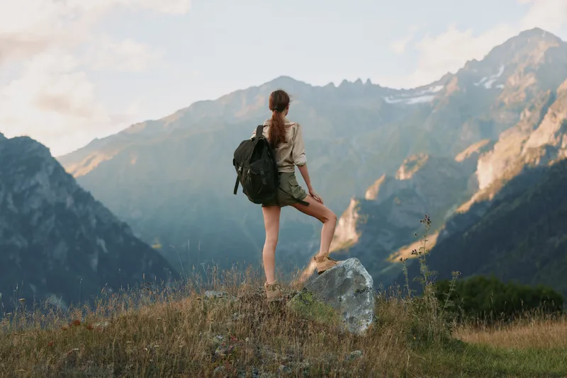 Young woman with a backpack stands on a rocky ledge, looking toward distant mountains, conveying a sense of adventure and quiet freedom.