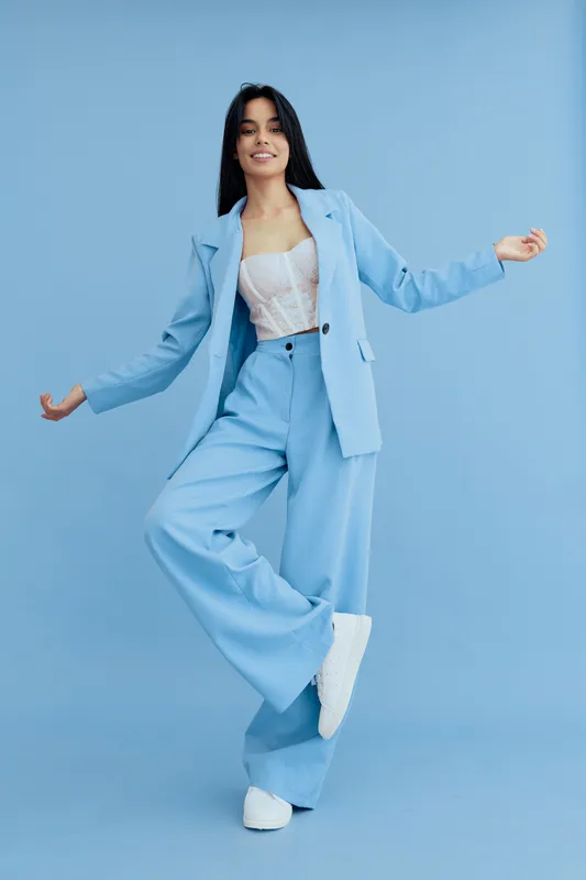 Young woman wearing stylish light blue suit and white lace top stands confidently with one leg raised against a solid blue background, exuding a fresh and dynamic vibe.