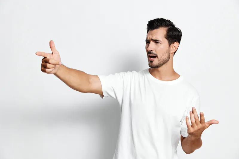 Young cheerful man gesturing with hands, expressing surprise and confusion, wearing a casual white t-shirt, standing against a plain light background. People lifestyle concept.
