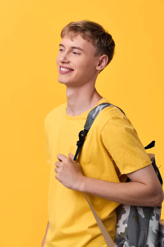 Smiling youth with a backpack, wearing a yellow tee, against a bright yellow backdrop, casual street style, ready for school or travel, positive mood, sunny color palette, everyday lifestyle scene
