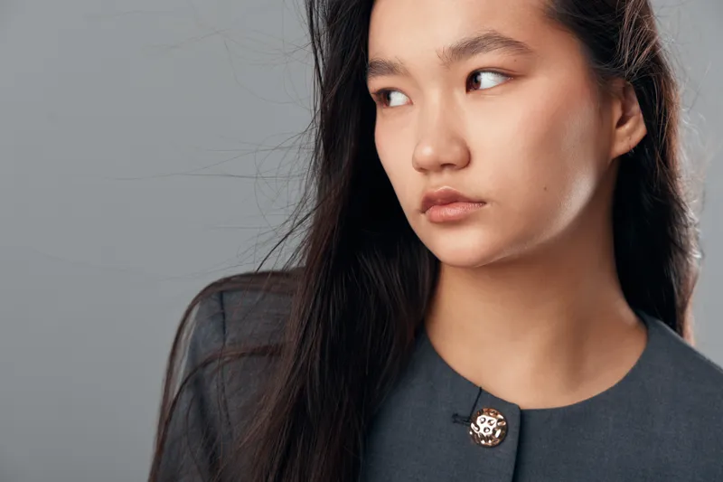 Young Asian woman with long black hair and natural makeup looking to the side in studio. Female model wearing dark gray top with simple button on plain gray background.