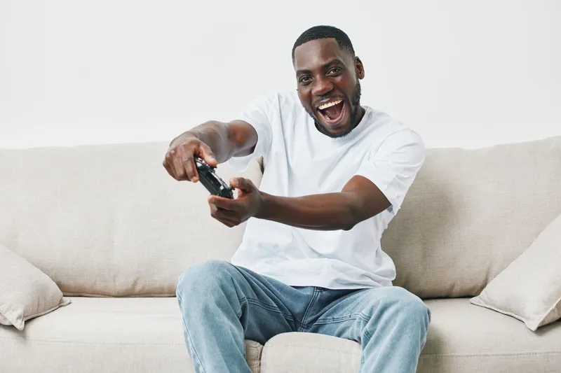 Happy young man playing video games on sofa, expressing excitement and joy with a gaming controller, casual outfit, home interior, energetic atmosphere