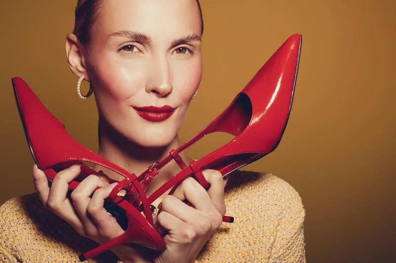 Close-up of confident woman holding red high heels near face, smiling softly. Warm lighting highlights her makeup and earrings on brown background. Concept of fashion and style.