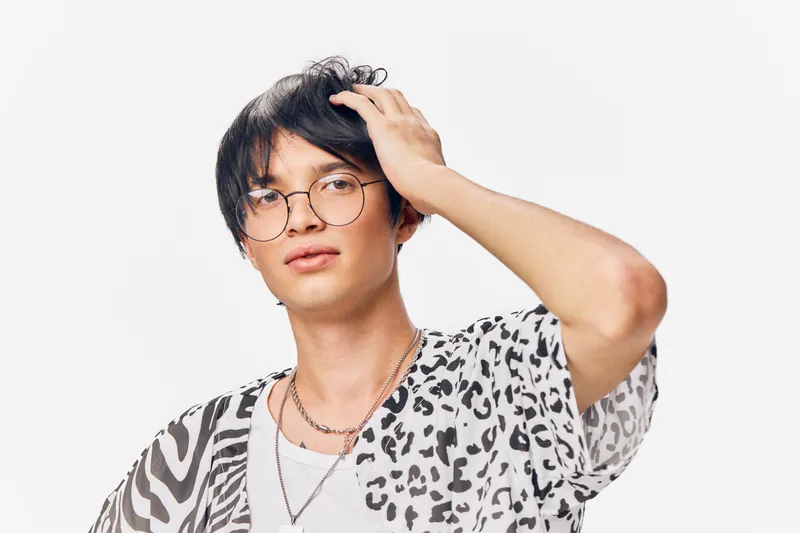 young man with glasses wearing patterned shirt and silver chains posing confidently against white background, stylish and modern casual look