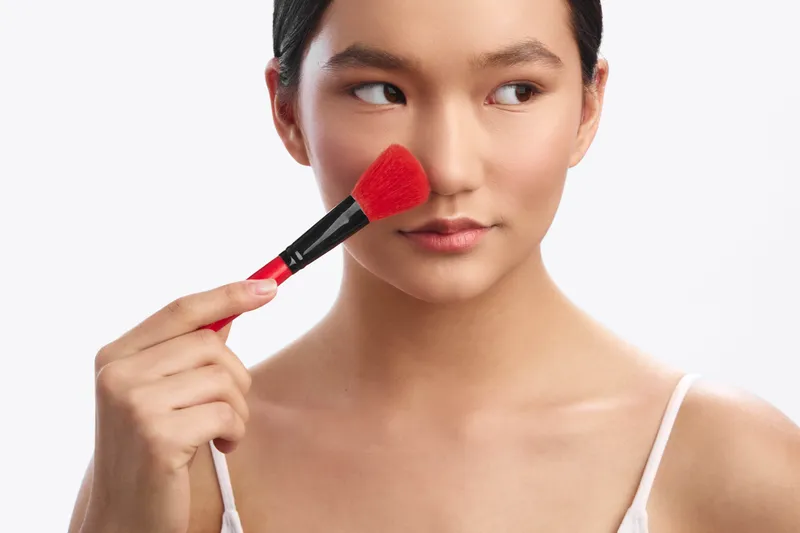 asian woman applies makeup with a soft brush in a clean studio setting, focused yet relaxed, highlighting beauty routine and self care in a calm and modern atmosphere.