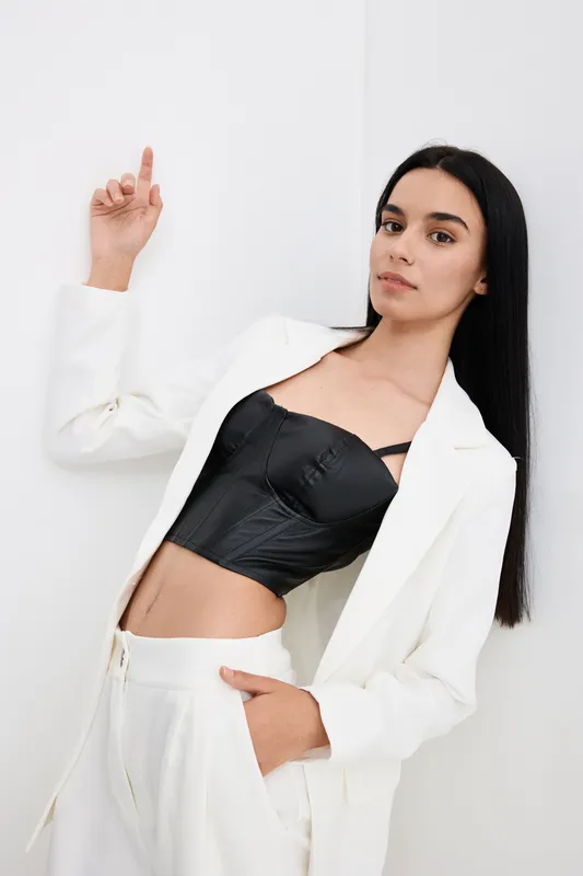 Young woman in white suit and black corset posing confidently against white background, showcasing modern fashion and elegant style with a minimalist vibe.
