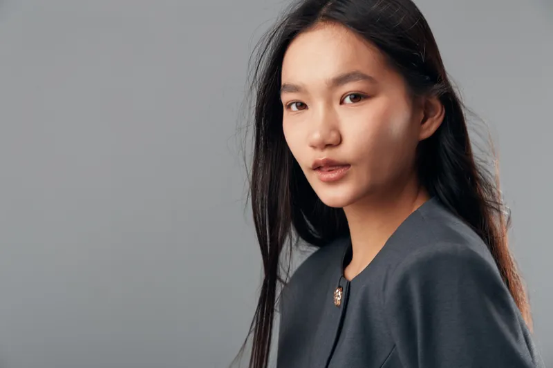 Young Asian woman in dark formal jacket with long hair posing against gray background for portrait, confidence, business style, professional look, elegance and subtle makeup.