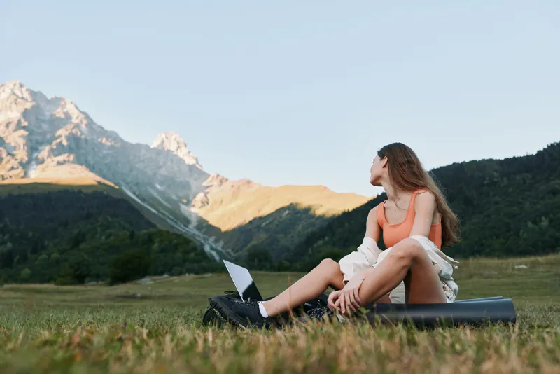 A relaxed outdoor scene shows a person sitting on the grass with a laptop, gazing toward distant mountains. Bright, natural light creates a calm, productive mood.