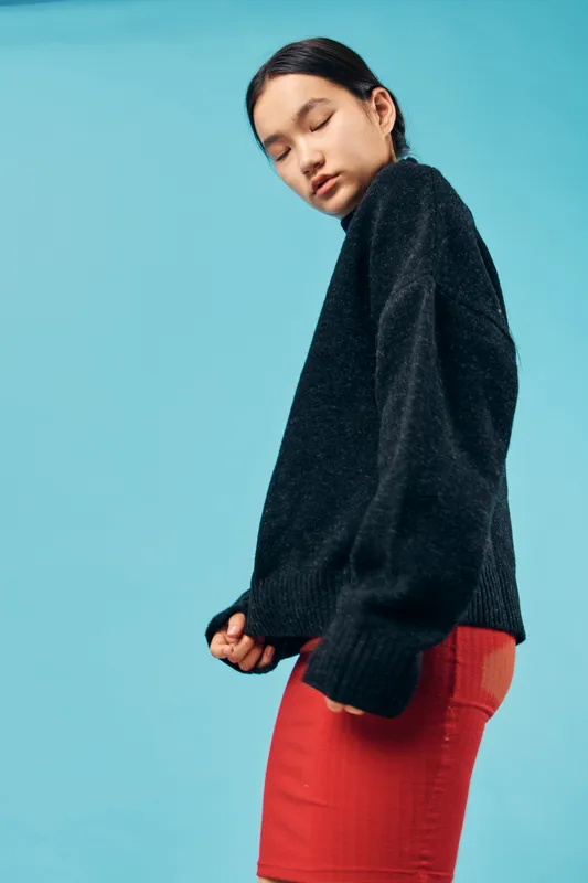 Young woman wearing black sweater and red skirt standing sideways with eyes closed on blue background. Fashion model in casual outfit with relaxed expression and minimal makeup indoor.