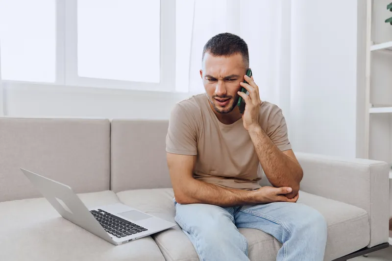 Concerned man talking on the phone while sitting on a couch, dressed casually, with a laptop nearby and a bright, airy room setting for modern lifestyle vibes.