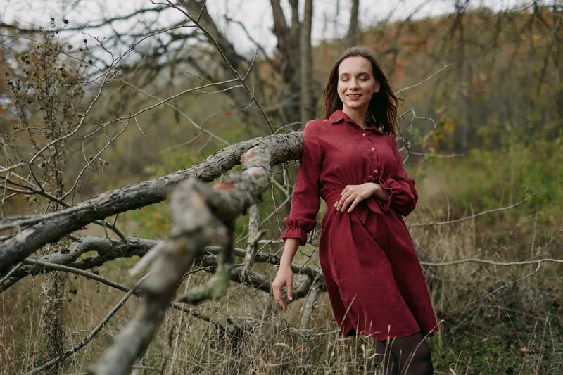 A candid portrait of a woman in a red dress leaning on a fallen tree in a quiet meadow, conveying natural authenticity and quiet confidence in rustic autumn light and a subtle smile.