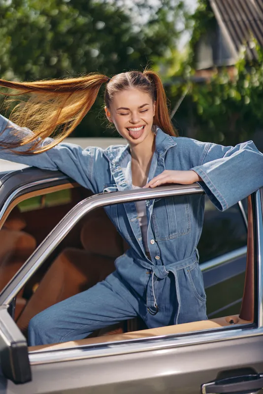 Cheerful woman in denim jumpsuit smiling from a vintage car, capturing the essence of a carefree summer day with vibrant greenery in the background.