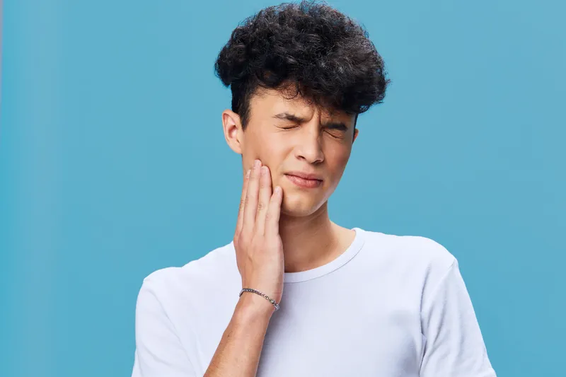 Young man in white t-shirt holding his jaw with painful expression, experiencing toothache, isolated on blue background with curly hair and casual style.