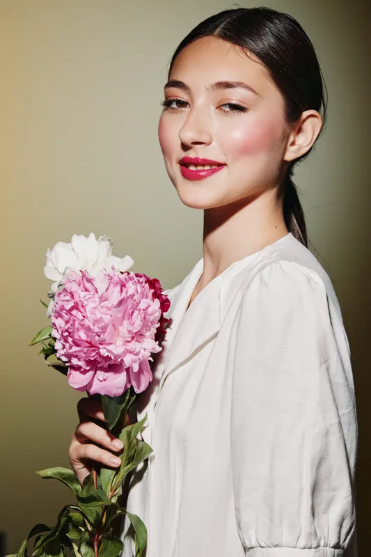 Young woman with glowing skin and bright lipstick holds a beautiful peony bouquet. Her hair is tied back, and she wears a simple white blouse in a soft studio setting.
