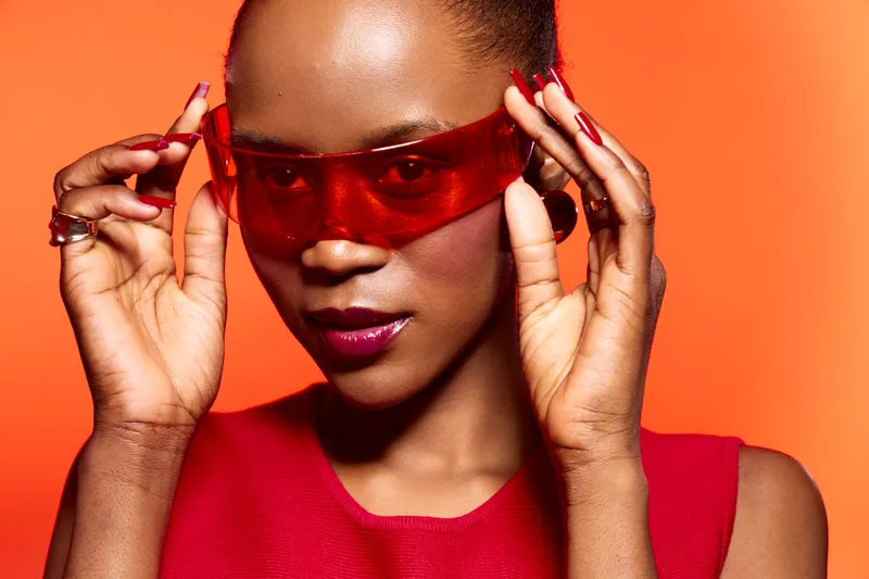 Portrait of confident African woman with dark skin wearing red sleeveless top and red sunglasses. Orange background highlights modern fashion and bold style concept with vibrant colors.