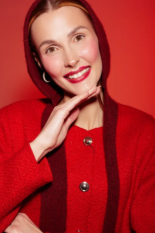 Smiling woman in red sweater and hood poses with hand near face against red background. Bright makeup and joyful expression create warm and festive mood in studio portrait.