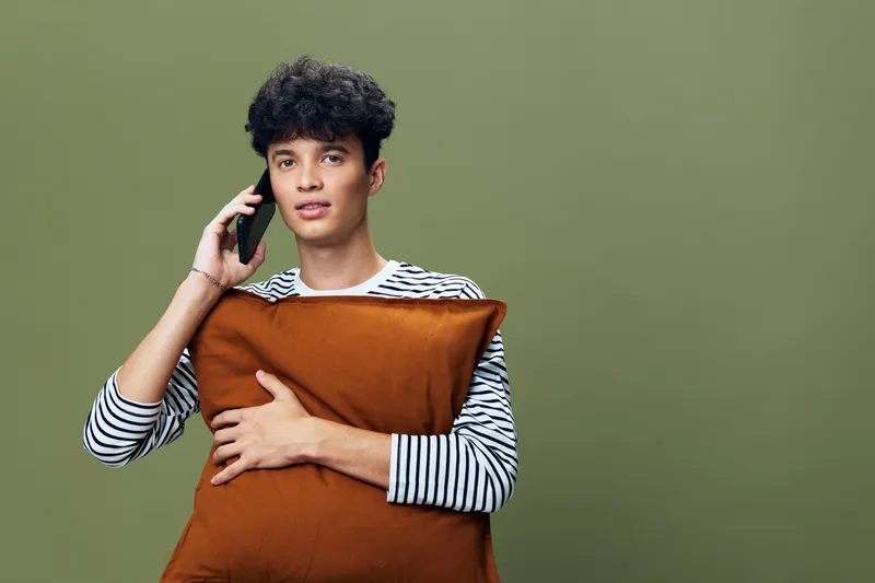 Young man talking on smartphone holding brown pillow, wearing striped shirt with curly hair, casual indoor setting and olive green background, relaxed expression.