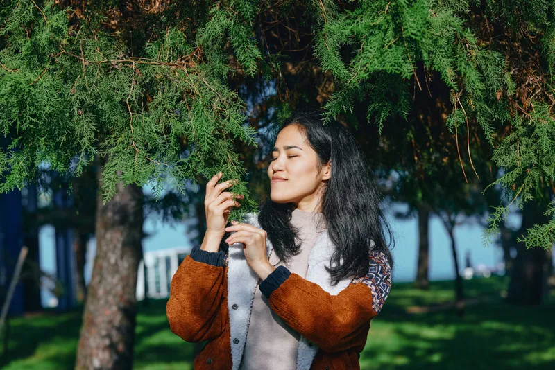 Woman beneath leafy trees in a sunny outdoor scene, wearing a warm brown cardigan and light scarf, eyes closed, savoring sunlight and breeze, serene mood, natural lifestyle moment.
