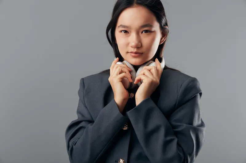 Young woman in dark gray blazer holding white headphones around her neck posing on gray background. Confident female with straight black hair and casual expression in studio shot.
