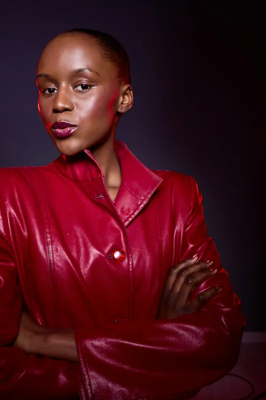 Confident dark skinned African woman wearing shiny red leather jacket poses with crossed arms against dark background. Concept of fashion, empowerment, beauty, and style.