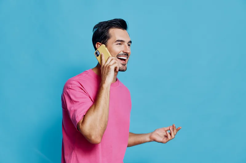 Smiling man with stylish haircut wearing pink t-shirt talking on mobile phone while standing against a bright blue background. People communication concept.