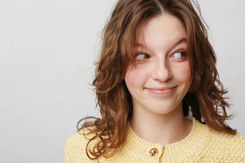 Young woman with wavy brown hair wearing yellow sweater looks sideways with a slight smile. Studio portrait on white background. Concept of casual style, natural beauty, and positive mood.