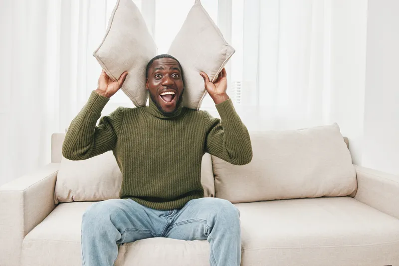 Happy man expressing joy with pillows in a modern living room, wearing cozy sweater and jeans, against a bright and airy background