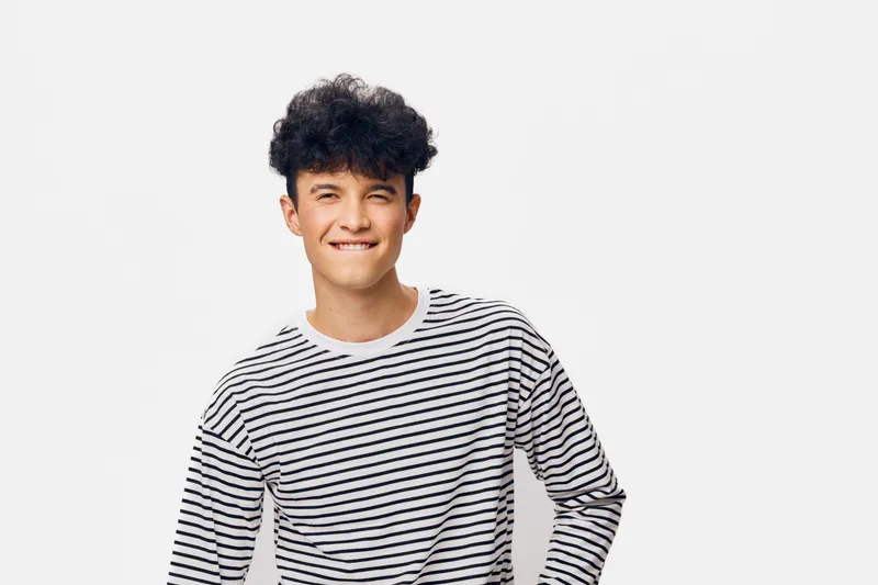 smiling man in casual striped shirt with curly hair standing against white background, expressing positive emotions and youthful energy in studio setting