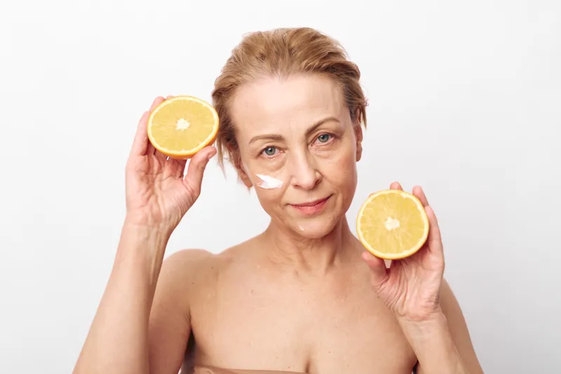 Mature woman with natural skin holds two halves of orange near her face with white cream on cheeks. Concept of skincare routine, healthy lifestyle, natural beauty, and vitamin C therapy.
