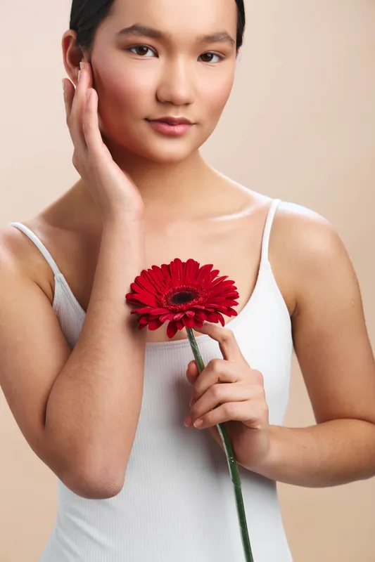 asian woman holds a bright red gerbera flower near her chest, touching her cheek with a gentle gaze, in a soft studio setting with warm neutral lighting.