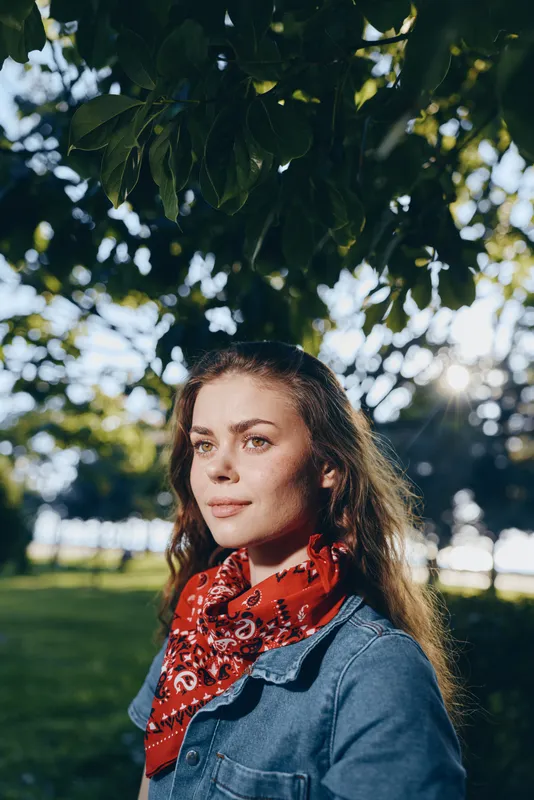 Lifestyle woman in modern boho-western denim street style wearing red bandana, standing outdoors with natural sunlight and green foliage in film color tone.