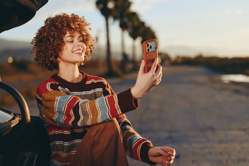 Woman lifestyle outdoors at sunset, smiling and holding a smartphone for a selfie near a car, curly hair and striped sweater, relaxed travel moment in golden hour, warm breeze today