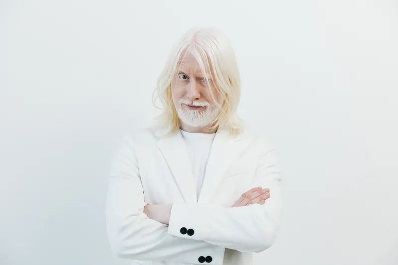 Уверенный мужчина с длинными седыми волосами и бородой в шикарном белом костюме позирует на светлом фоне, излучая своим выражением лица игривую, но в то же время изысканную атмосферу