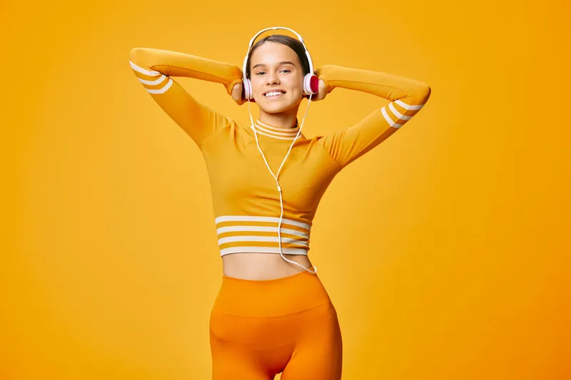 Woman in orange fitness outfit posing with hands behind head, headphones on, smiling against a bright orange background; a dynamic sport scene emphasizing wellness and energy.
