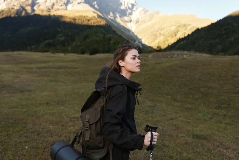 A solo female hiker stands in a grassy valley with a backpack and trekking pole, gazing toward distant mountain peaks under a clear sky, embodying adventure, resilience, and exploration.