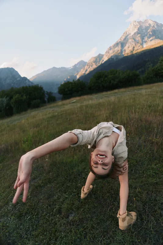 Young woman performs a backbend in a grassy meadow with distant mountains, capturing a playful, adventurous moment outdoors.