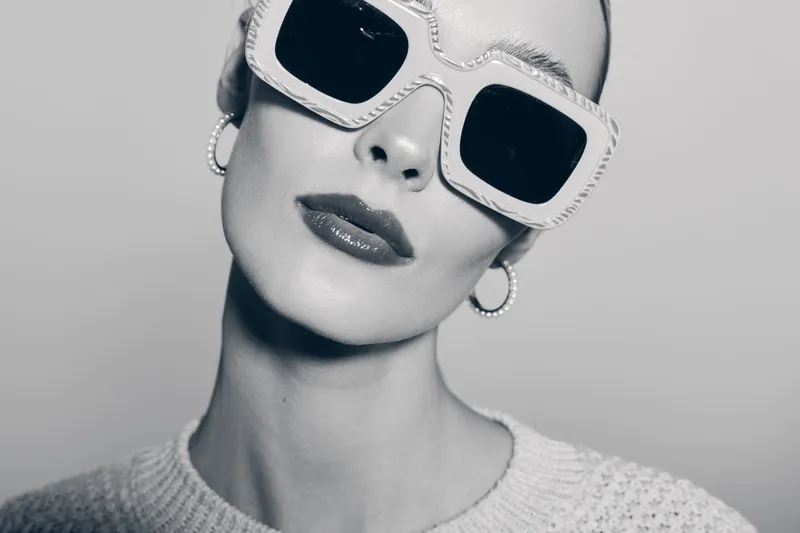 Stylish white woman with pale skin wears large sunglasses and hoop earrings. Fashion portrait in monochrome with elegant makeup and knitted sweater.